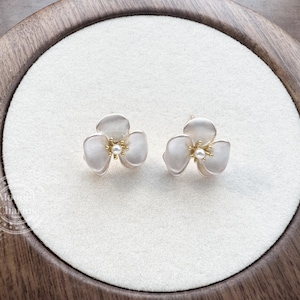 Pode incluir: Um par de brincos de tarracha em formato de flores de tr&ecirc;s p&eacute;talas com acabamento branco fosco e um pequeno detalhe de p&eacute;rola central. Os brincos apresentam estames de metal dourado no centro de cada flor, criando um contraste delicado.