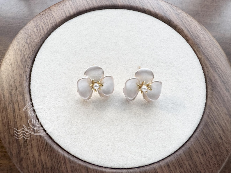 Pode incluir: Um par de brincos de tarracha em formato de flores de tr&ecirc;s p&eacute;talas com acabamento branco fosco e um pequeno detalhe de p&eacute;rola central. Os brincos apresentam estames de metal dourado no centro de cada flor, criando um contraste delicado.