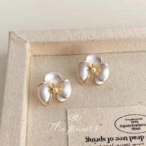 Pode incluir: Um par de brincos de tarracha em formato de flor de tr&ecirc;s p&eacute;talas com acabamento branco perolado. Cada brinco apresenta um estame central em tom dourado com uma pequena conta branca, exibidos sobre uma superf&iacute;cie de tecido bege.