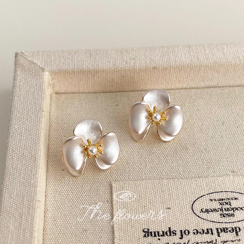 Pode incluir: Um par de brincos de tarracha em formato de flor de tr&ecirc;s p&eacute;talas com acabamento branco perolado. Cada brinco apresenta um estame central em tom dourado com uma pequena conta branca, exibidos sobre uma superf&iacute;cie de tecido bege.