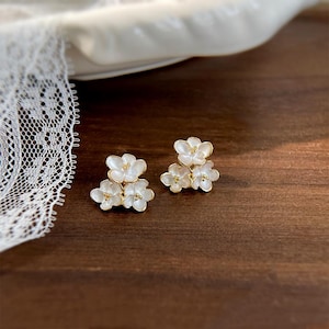 May include: A pair of gold-tone earrings with three white flower charms each. The flowers have a pearl-like finish and are accented with tiny gold dots.