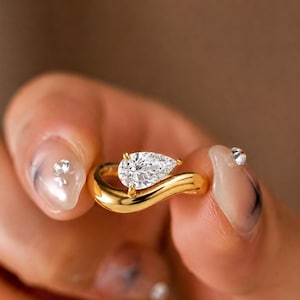 May include: A gold ring featuring a single pear-shaped clear gemstone set on a smooth, curved band. The metal has a polished finish, and the stone is secured by small prongs, creating a sleek, continuous design that wraps around the finger.