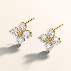 May include: A pair of gold stud earrings, each featuring four round, clear, faceted stones arranged in a clover-like pattern with a central star-shaped gold accent. The text MoonlitCharms is visible near the left earring. The posts are gold.