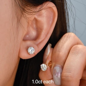 Puede incluir: Primer plano de dos pendientes de oro, cada uno con un diamante redondo de talla brillante. El texto "1.0ct each" es visible. Los pendientes están engastados en un engaste de oro.