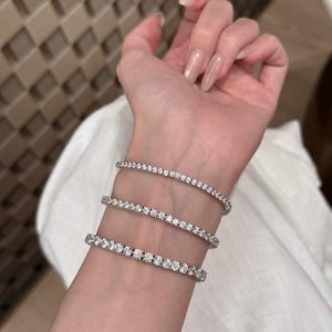 May include: Three silver-toned tennis bracelets featuring a continuous row of small, clear, round-cut stones. The bracelets are worn stacked on the wrist, showcasing a metallic finish and a uniform, sparkling design across each individual piece.