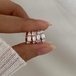 Può includere: Primo piano di tre paia di orecchini a cerchio. Due paia sono color argento e uno color oro. Ogni orecchino presenta una fila di piccole pietre scintillanti, rettangolari e a forma di pera. Gli orecchini sono tenuti tra le dita.