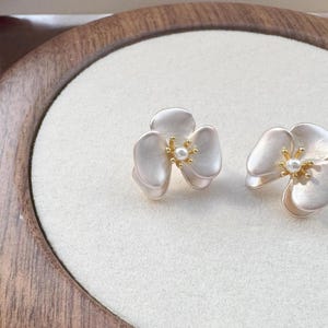 Pode incluir: Um par de brincos em forma de flor de tr&ecirc;s p&eacute;talas com acabamento mate em branco sujo, apresentando um estame central dourado com uma pequena p&eacute;rola branca. O texto Moonlit Charms &eacute; vis&iacute;vel no canto inferior esquerdo da imagem.