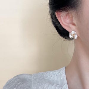 Pode incluir: Um brinco de pino em forma de flor de tr&ecirc;s p&eacute;talas com p&eacute;talas brancas iridescentes e lisas, e um pequeno centro texturizado de cor dourada. O brinco &eacute; usado no l&oacute;bulo da orelha, exibindo o design floral contra um fundo neutro.