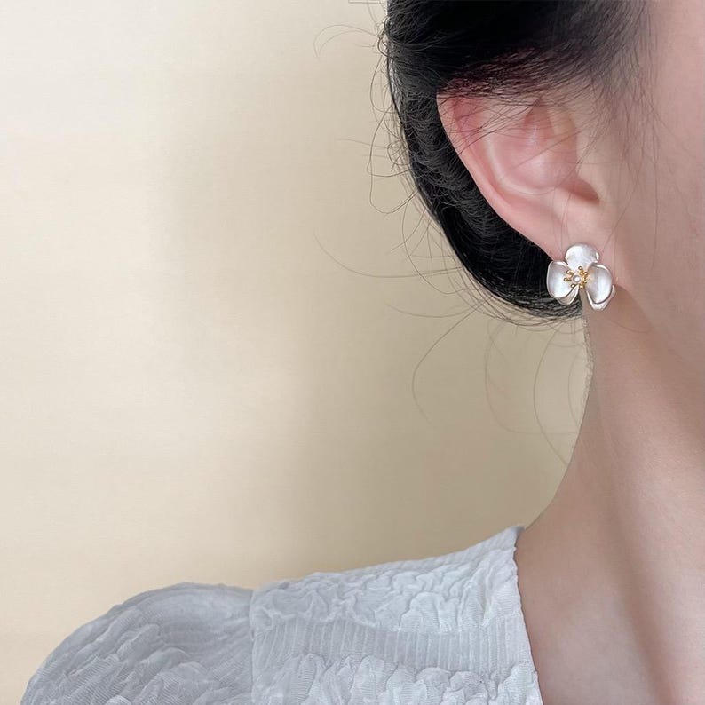 Pode incluir: Um brinco de pino em forma de flor de tr&ecirc;s p&eacute;talas com p&eacute;talas brancas iridescentes e lisas, e um pequeno centro texturizado de cor dourada. O brinco &eacute; usado no l&oacute;bulo da orelha, exibindo o design floral contra um fundo neutro.