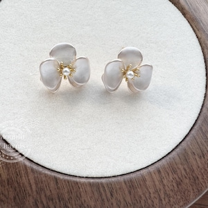 Pode incluir: Um par de brincos de tarracha em forma de flores de tr&ecirc;s p&eacute;talas. Cada brinco apresenta p&eacute;talas brancas foscas com um estame central em tom dourado rematado por uma pequena p&eacute;rola branca. Exibidos sobre uma superf&iacute;cie circular de tecido branco.