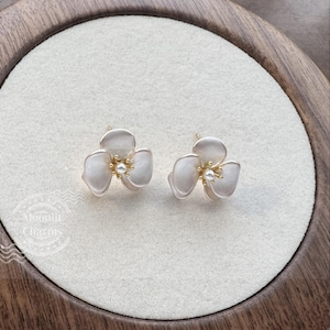 Pode incluir: Um par de brincos em forma de flor de tr&ecirc;s p&eacute;talas com um acabamento mate em branco sujo. Cada brinco apresenta um pequeno estame central dourado rematado com uma min&uacute;scula p&eacute;rola branca. O texto Moonlit Charms &eacute; vis&iacute;vel no canto.