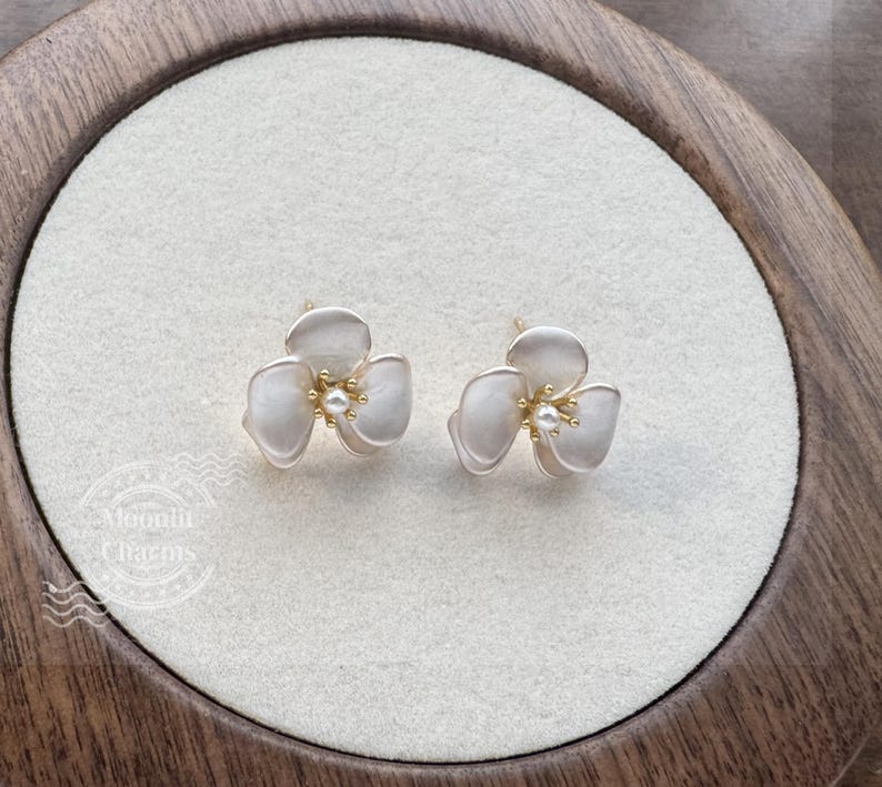 Pode incluir: Um par de brincos em forma de flor de tr&ecirc;s p&eacute;talas com um acabamento mate em branco sujo. Cada brinco apresenta um pequeno estame central dourado rematado com uma min&uacute;scula p&eacute;rola branca. O texto Moonlit Charms &eacute; vis&iacute;vel no canto.