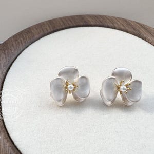 Pode incluir: Um par de brincos de tarracha em forma de flores de tr&ecirc;s p&eacute;talas. Cada brinco apresenta p&eacute;talas brancas foscas com um centro em tom dourado e um pequeno detalhe de p&eacute;rola branca. O design &eacute; sim&eacute;trico, exibindo uma est&eacute;tica floral delicada.