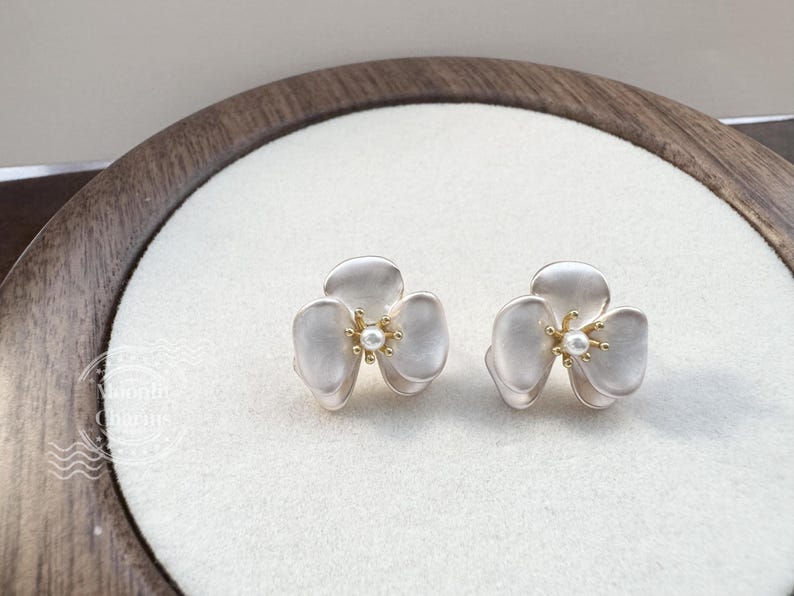 Pode incluir: Um par de brincos de tarracha em forma de flores de tr&ecirc;s p&eacute;talas. Cada brinco apresenta p&eacute;talas brancas foscas com um centro em tom dourado e um pequeno detalhe de p&eacute;rola branca. O design &eacute; sim&eacute;trico, exibindo uma est&eacute;tica floral delicada.