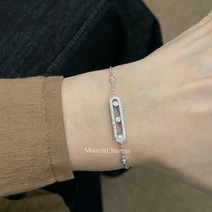 May include: A delicate silver bracelet with a rectangular centerpiece adorned with three clear, round gemstones. The bracelet is attached to a fine chain with additional small gemstones. The text "MoonlitCharms" is visible.