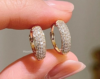 Puede incluir: Un par de pendientes de aro dorados, incrustados con pequeñas piedras brillantes. Los pendientes están sostenidos por los dedos, mostrando su diseño y la forma en que capturan la luz. Son una pieza de joyería.