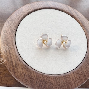Pode incluir: Um par de brincos de tarracha em forma de flores de tr&ecirc;s p&eacute;talas com um acabamento branco suave, centros em tom dourado e um pequeno detalhe de p&eacute;rola central. O texto Moonlit Charms &eacute; vis&iacute;vel no canto inferior esquerdo da imagem.