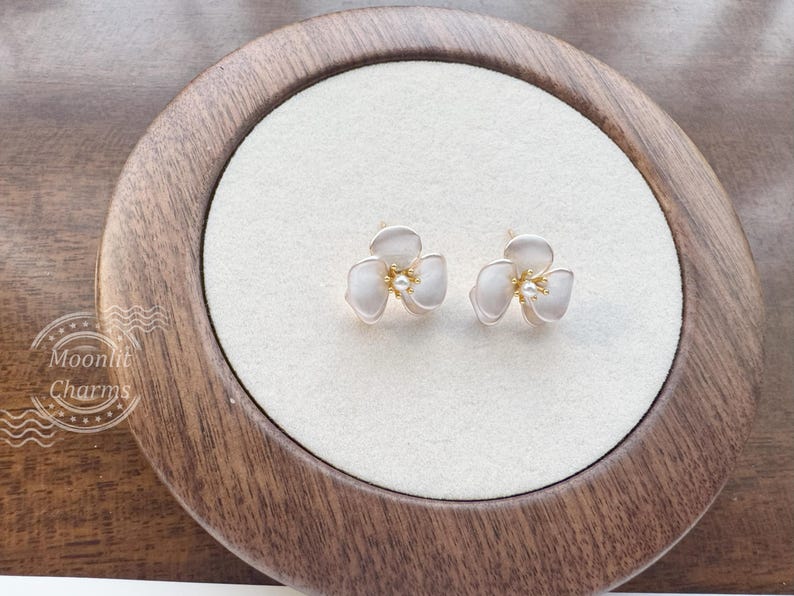 Pode incluir: Um par de brincos de tarracha em forma de flores de tr&ecirc;s p&eacute;talas com um acabamento branco suave, centros em tom dourado e um pequeno detalhe de p&eacute;rola central. O texto Moonlit Charms &eacute; vis&iacute;vel no canto inferior esquerdo da imagem.