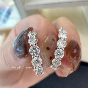 May include: A pair of silver-toned hoop earrings featuring a row of five round, clear, faceted stones set in each piece. The text MoonlitCharms is printed in white on the left side of the image. The earrings have a polished metallic finish.