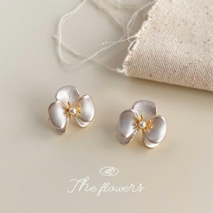 Pode incluir: Um par de brincos em forma de flores de tr&ecirc;s p&eacute;talas com acabamento branco perolado, centros dourados e uma pequena conta branca no meio. O texto "The flowers" est&aacute; impresso na parte inferior da imagem em fonte cursiva.