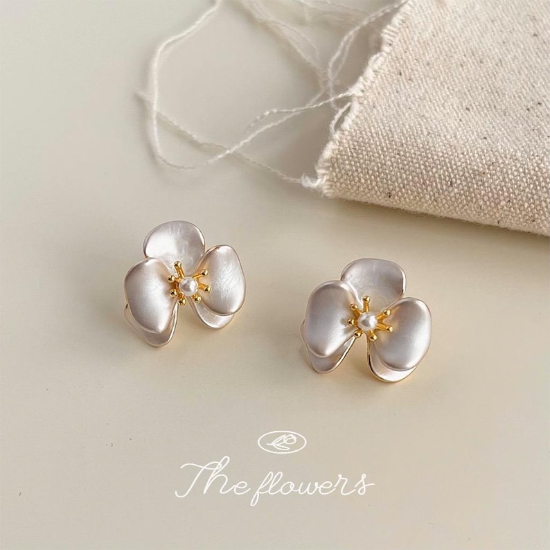 Pode incluir: Um par de brincos em forma de flores de tr&ecirc;s p&eacute;talas com acabamento branco perolado, centros dourados e uma pequena conta branca no meio. O texto "The flowers" est&aacute; impresso na parte inferior da imagem em fonte cursiva.