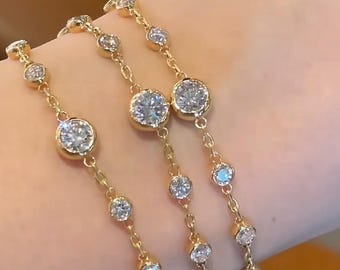 Puede incluir: Tres pulseras doradas con piedras preciosas redondas y transparentes. Cada pulsera presenta una delicada cadena con una serie de pequeñas gemas y piedras más grandes engarzadas. Las pulseras se muestran en un brazo.
