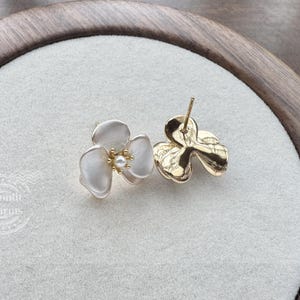 Pode incluir: Um par de brincos de tarracha com design de flor de tr&ecirc;s p&eacute;talas em acabamento branco fosco e uma pequena p&eacute;rola central. Um brinco mostra a frente, o outro o verso em metal dourado com pino. O texto Moonlit Charms est&aacute; vis&iacute;vel.