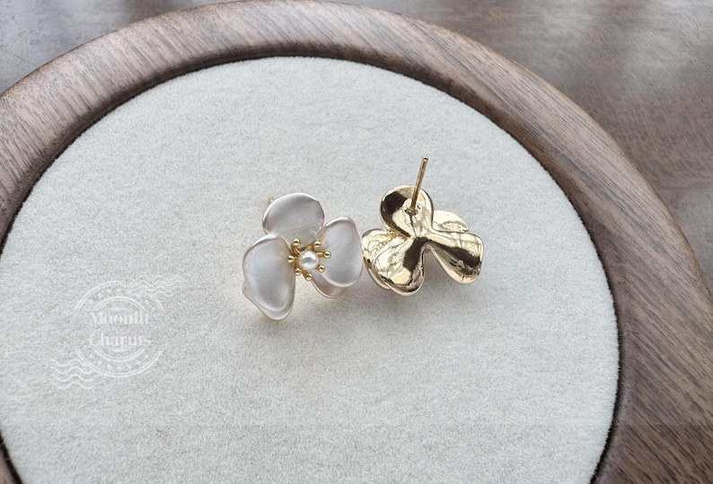 Pode incluir: Um par de brincos de tarracha com design de flor de tr&ecirc;s p&eacute;talas em acabamento branco fosco e uma pequena p&eacute;rola central. Um brinco mostra a frente, o outro o verso em metal dourado com pino. O texto Moonlit Charms est&aacute; vis&iacute;vel.