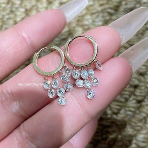 May include: Gold hoop earrings with dangling clear crystals. The earrings have a simple, circular design with multiple small, round crystals hanging from the bottom. The text "MoonlitCharm" is visible on the image.