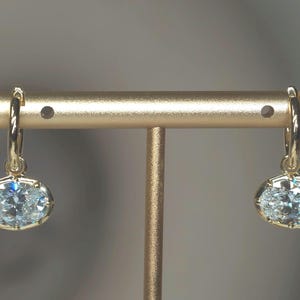 May include: A pair of gold hoop earrings, each featuring a single oval-cut clear gemstone suspended from the bottom. The stones are set in gold prongs, reflecting light with a bright, multifaceted sparkle against the polished gold metal hoops.