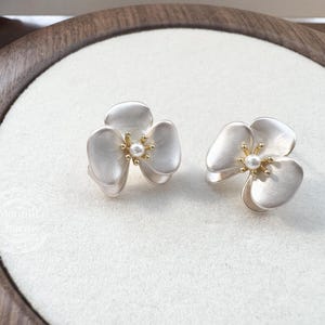 Pode incluir: Um par de brincos de tarracha em forma de flor de tr&ecirc;s p&eacute;talas com acabamento em prata fosca. Cada flor apresenta um estame central dourado com uma pequena p&eacute;rola branca. Design delicado com p&eacute;talas sobrepostas.
