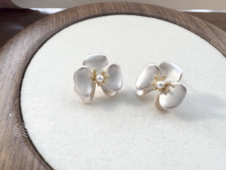 Pode incluir: Um par de brincos de tarracha em forma de flor de tr&ecirc;s p&eacute;talas com acabamento em prata fosca. Cada flor apresenta um estame central dourado com uma pequena p&eacute;rola branca. Design delicado com p&eacute;talas sobrepostas.