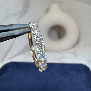 May include: A gold ring set with a line of clear, sparkling gemstones. The ring is held by tweezers against a blurred background. The gemstones are round and evenly spaced along the band, creating a continuous line of brilliance.