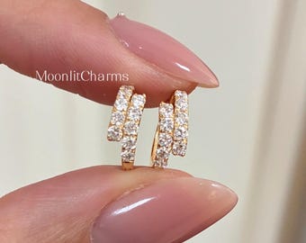 Puede incluir: Un par de pendientes de aro dorados con tres filas de pequeñas piedras preciosas transparentes. Los pendientes se sostienen entre dos dedos. El texto "MoonlitCharms" es visible en el fondo.