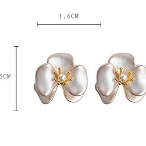 Pode incluir: Um par de brincos em forma de flor com p&eacute;talas brancas e um estame central dourado com uma pequena p&eacute;rola branca. Os brincos medem 1,6 cm de largura e 1,5 cm de altura, conforme indicado pelas linhas de medi&ccedil;&atilde;o na imagem.
