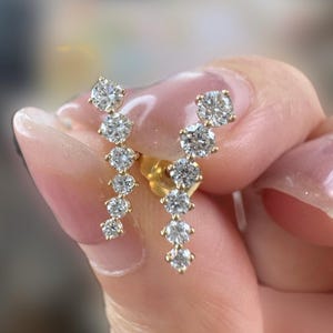 Puede incluir: Un par de pendientes de oro con un diseño graduado de diamantes redondos brillantes. Los pendientes están engastados en metal de oro amarillo y se muestran sobre un fondo borroso. Los pendientes son una joya.