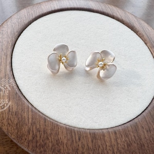 Pode incluir: Um par de brincos de tarracha em forma de flor de tr&ecirc;s p&eacute;talas com acabamento branco fosco. Cada brinco apresenta um centro dourado com um pequeno detalhe de p&eacute;rola branca. Os brincos est&atilde;o expostos numa base circular branca.