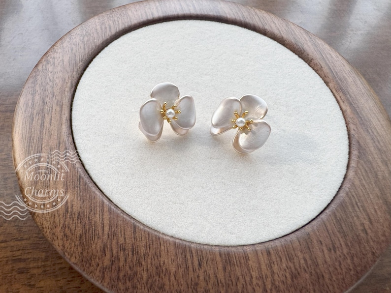 Pode incluir: Um par de brincos de tarracha em forma de flor de tr&ecirc;s p&eacute;talas com acabamento branco fosco. Cada brinco apresenta um centro dourado com um pequeno detalhe de p&eacute;rola branca. Os brincos est&atilde;o expostos numa base circular branca.