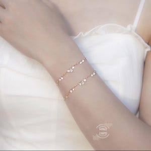 May include: Two delicate gold bracelets, each adorned with sparkling clear crystals, are worn on an arm. The bracelets are layered, creating a subtle yet elegant look. The overall aesthetic is minimalist and refined, perfect for adding a touch of sparkle.