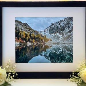 A framed photograph of a mountain lake with snow-capped mountains in the background. The lake is reflecting the mountains and the sky. The trees on the mountainside are a mix of green and yellow. The frame is black and has a white mat.