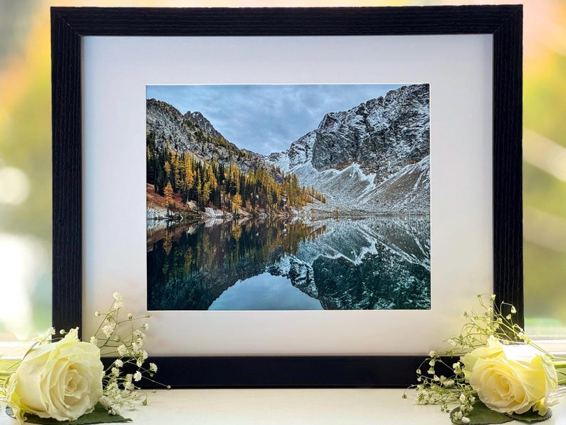 A framed photograph of a mountain lake with snow-capped mountains in the background. The lake is reflecting the mountains and the sky. The trees on the mountainside are a mix of green and yellow. The frame is black and has a white mat.
