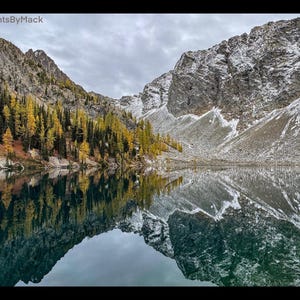 May include: A mountain lake with a reflection of the surrounding mountains and trees. The water is still and the sky is cloudy. The mountains are covered in snow and the trees are a mix of green and yellow.