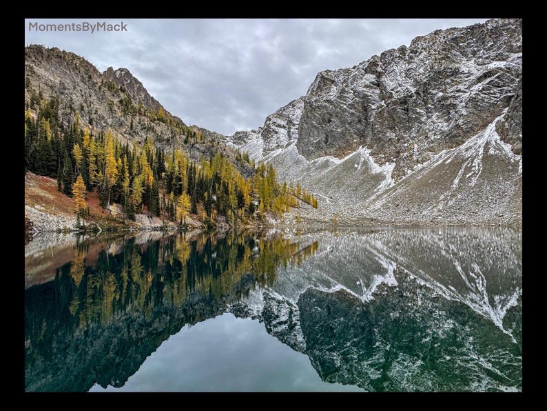 May include: A mountain lake with a reflection of the surrounding mountains and trees. The water is still and the sky is cloudy. The mountains are covered in snow and the trees are a mix of green and yellow.