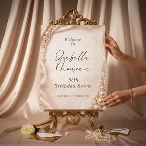 May include: A rectangular welcome sign featuring a cream-colored background with a pearl border, displayed on an ornate gold easel. The text reads: Welcome To Isabella Thonpe & 30th Birthday Soirée, Saturday, October 12th. The design includes soft fabric folds.