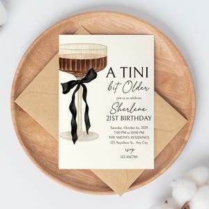 Puede incluir: Una tarjeta de invitaci&oacute;n con el texto "A Tini bit Older" y "Sherlene 21st Birthday". La tarjeta presenta una ilustraci&oacute;n de acuarela de una copa de c&oacute;ctel con una cinta negra. La tarjeta est&aacute; sobre un plato de madera con un sobre.