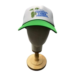 May include: White trucker hat with a green brim. The hat features the text "LAGUNITAS CONTENTS UNDER FRESHER" in blue and a green graphic of hops. The hat is displayed on a mannequin head.