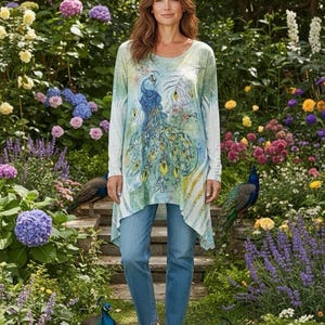 Puede incluir: Una mujer con una túnica de manga larga con un diseño de pavo real, combinada con vaqueros azules y zapatillas blancas, posa en un jardín. La túnica presenta una ilustración de pavo real azul y verde. El jardín está lleno de flores coloridas y varios pavos reales.