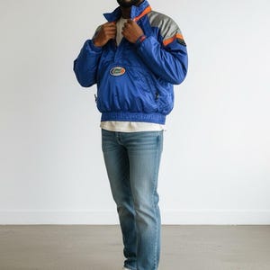 May include: A blue and gray windbreaker jacket with orange accents and a logo patch. The jacket is paired with blue jeans and white sneakers. The outfit is casual and sporty.