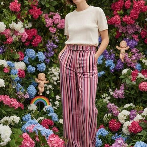 May include: A woman wearing a cream-colored short-sleeved top and striped pants with a belt. The pants have vertical stripes in shades of pink, black, and white. The background is a lush garden of colorful flowers, including pink, blue, and white hydrangeas.