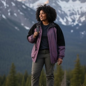 May include: A person wearing a purple and black jacket, dark green pants, and hiking boots stands in front of a mountain range. The jacket has a black collar and sleeves, with a purple body. The person is smiling.